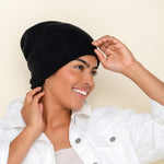 A smiling person wearing a black, 6-ply ribbed knit cashmere Watchcap paired with a white denim jacket. 